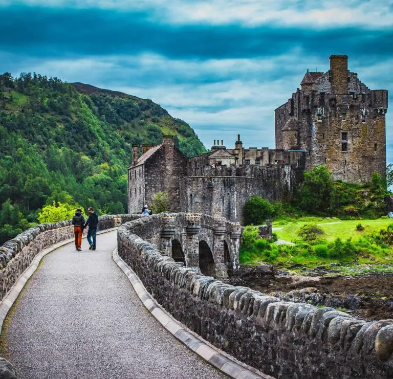 Tourists walking across the stone bridge towards Eilean Donan Castle surrounded by lush green hills in the Scottish Highlands, Scotland