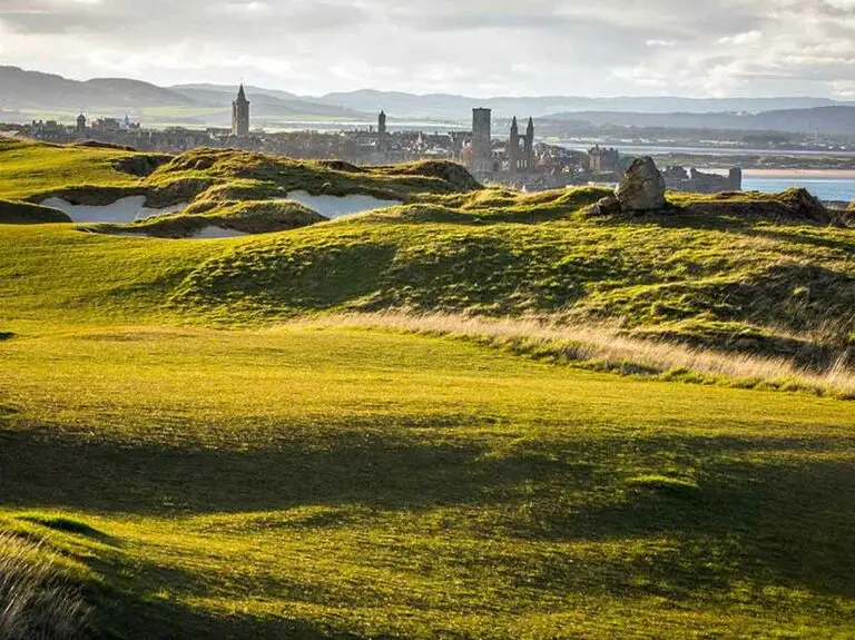 Scenic view of Muirfield Golf Course in Scotland with rolling green fairways and distant historic architecture under a cloudy sky