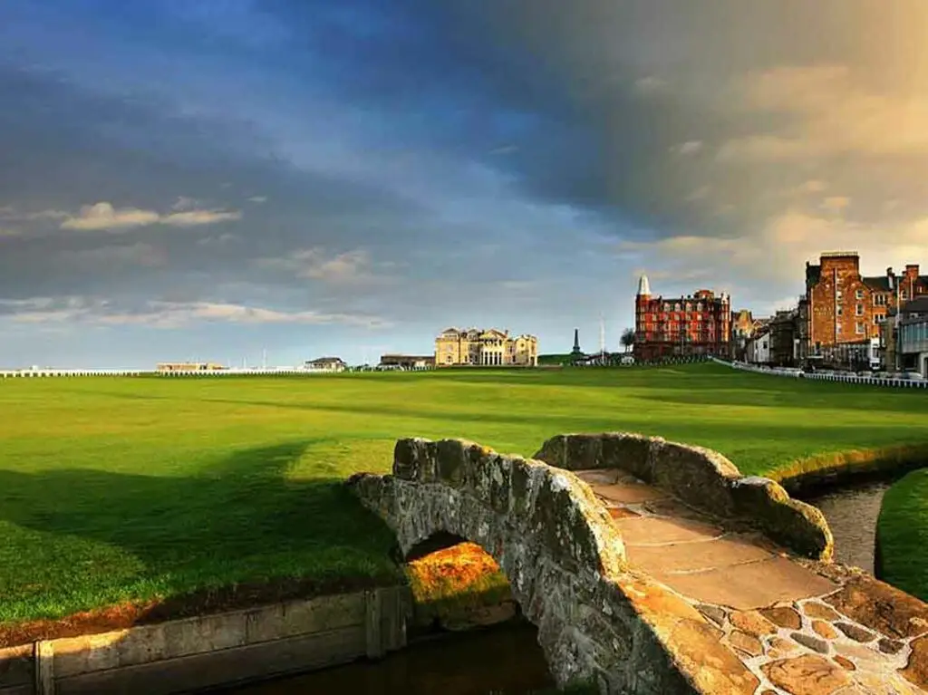 Historic Swilcan Bridge on the Old Course golf course in St Andrews, Scotland, with the Royal and Ancient Clubhouse and town buildings in the background