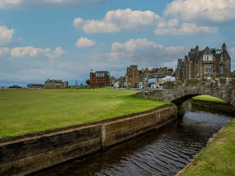 Stone bridge over the Swilcan Burn on the Old Course at St Andrews, Scotland, with historic buildings and golf links under a cloudy sky