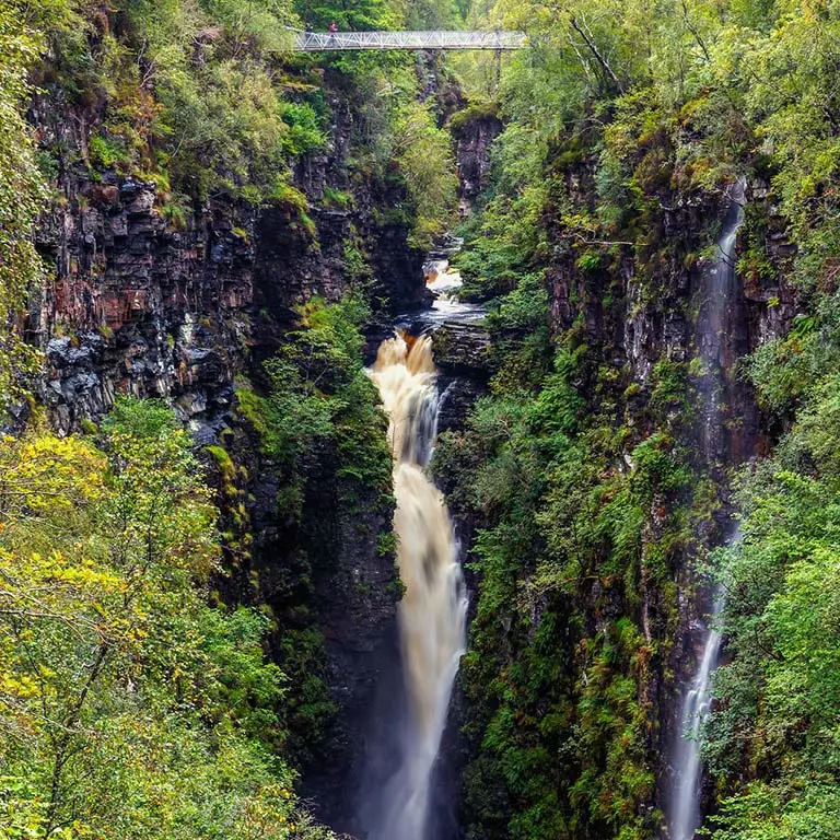 Waterfall cascading through the lush green cliffs of Corrieshalloch Gorge in Scotland with a viewing bridge above, a popular Scottish tourist attraction