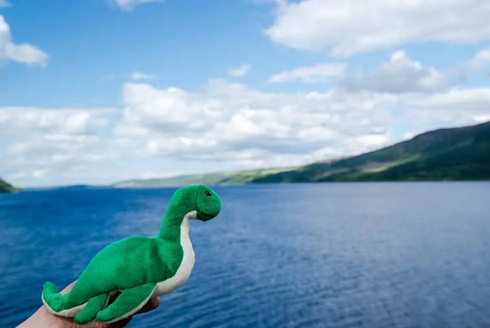 Toy Nessie monster held in front of Loch Ness with scenic Scottish Highlands landscape and blue sky, Scotland tourism attraction