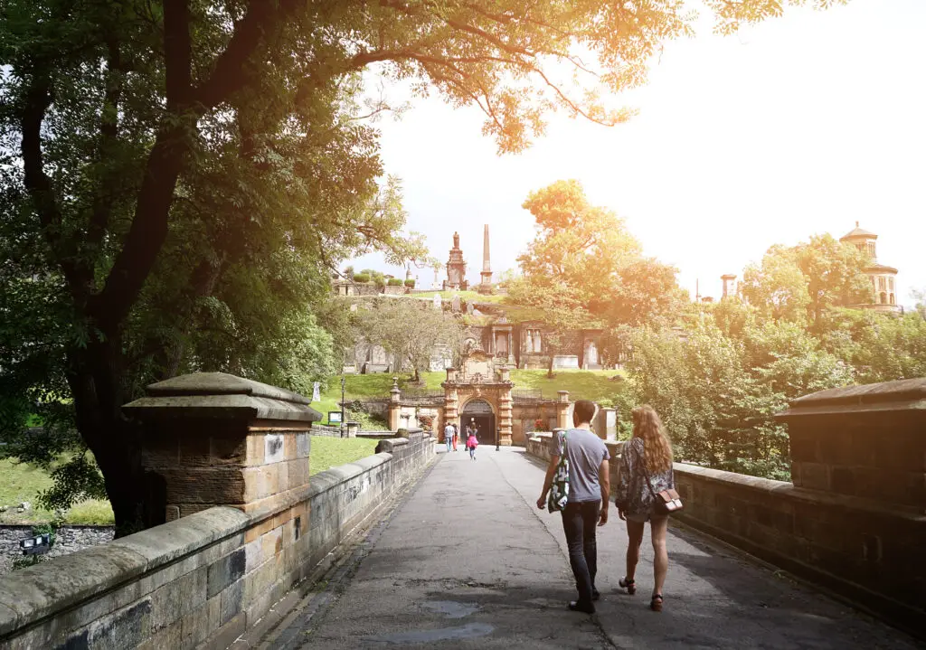 Tourists walking across a stone bridge toward the historic Glasgow Necropolis surrounded by greenery and sunshine in Glasgow, Scotland
