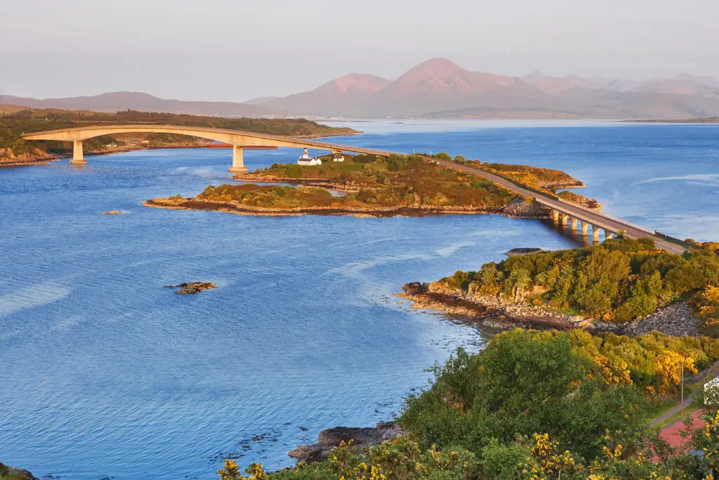 Scenic view of the Isle of Skye Bridge in Scotland with blue waters, lush green hills, and distant mountains at sunset