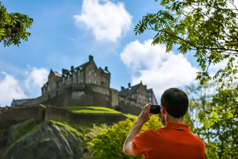 Tourist taking a photo of Edinburgh Castle in Scotland on a sunny day surrounded by greenery