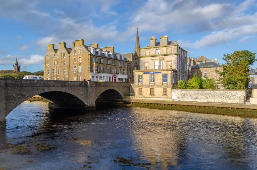 Historic stone bridge and traditional buildings along the river in Wick, Scotland, under a bright blue sky