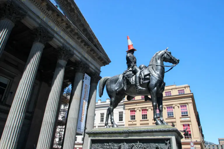 Equestrian statue in front of the Gallery of Modern Art in Glasgow, Scotland, with a traffic cone on the rider’s head under a bright blue sky
