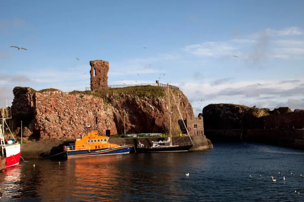 Boats docked at Dunbar Harbour with historic red sandstone ruins and cliffs in East Lothian, Scotland, under a bright blue sky