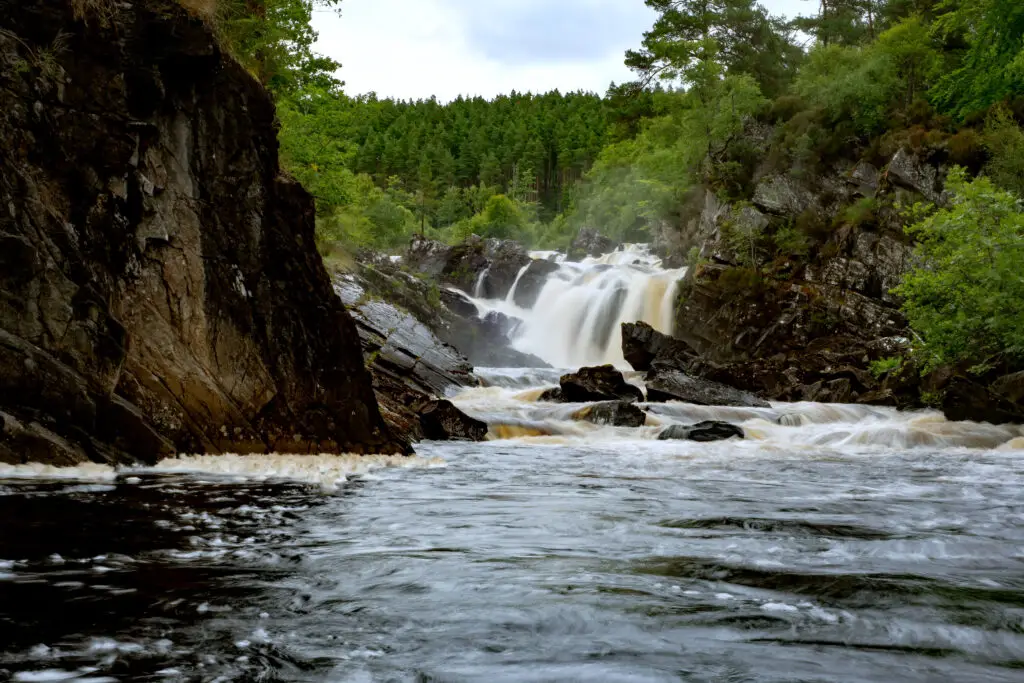 Rogie Falls in Scotland surrounded by lush green forest and rocky cliffs with cascading water flowing through the gorge, a popular Scottish Highlands tourist attraction