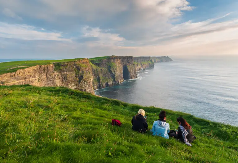 Three girls sitting on green grass near the edge of the Cliffs of Moher in Ireland overlooking the Atlantic Ocean on a scenic day