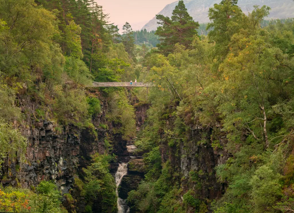 Pedestrian suspension bridge over Corrieshalloch Gorge with cascading waterfall surrounded by lush green forest in Scotland