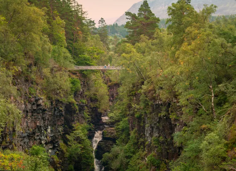 Pedestrian suspension bridge over Corrieshalloch Gorge with cascading waterfall surrounded by lush green forest in Scotland