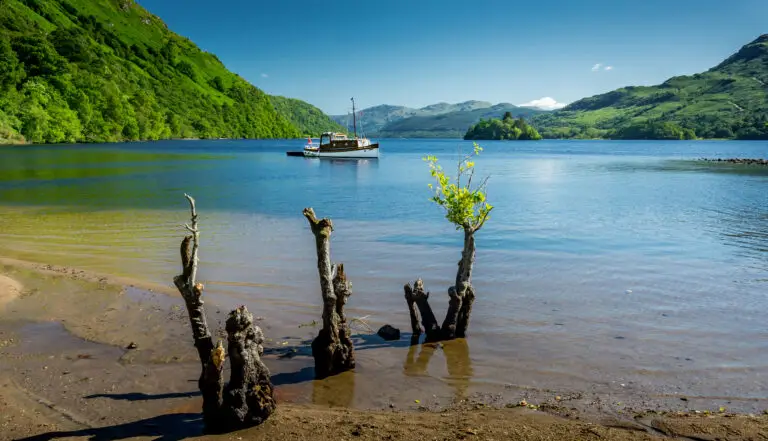 Scenic view of Loch Lomond in Scotland with a small boat on calm blue water surrounded by green hills and a sandy shoreline, popular destination for Scottish tours and nature travel