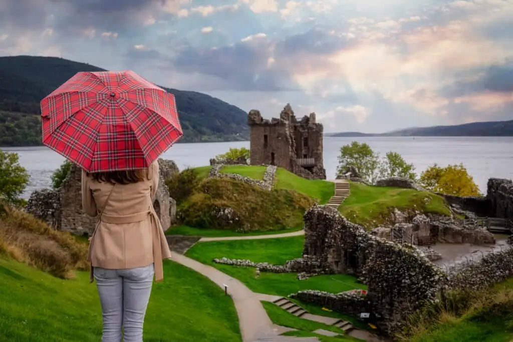 Tourist holding a red tartan umbrella overlooking Urquhart Castle ruins on the shores of Loch Ness in the Scottish Highlands, Scotland