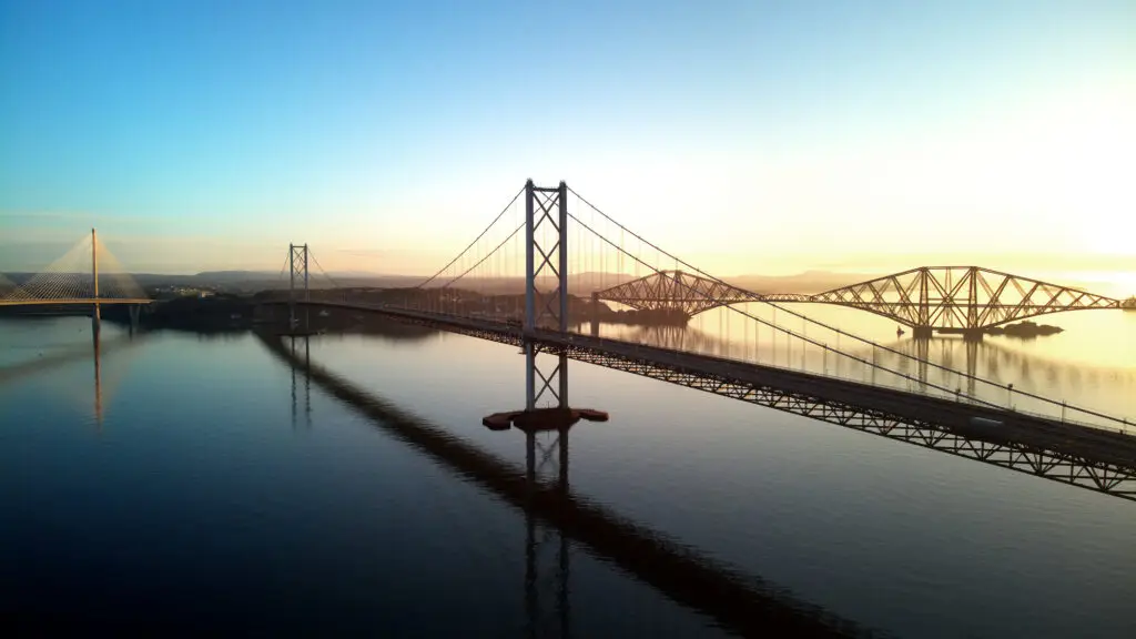 Sunrise view of the Forth Bridges spanning the Firth of Forth near Edinburgh, Scotland, featuring the iconic Forth Rail Bridge, Forth Road Bridge, and Queensferry Crossing reflecting on calm waters.