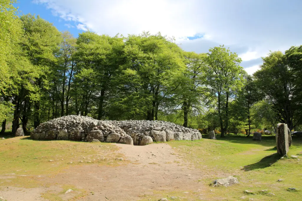 Ancient stone cairn and standing stones at Balnuaran of Clava in Strathnairn, Scotland surrounded by green trees and blue sky, a historic Scottish tourist attraction near Inverness