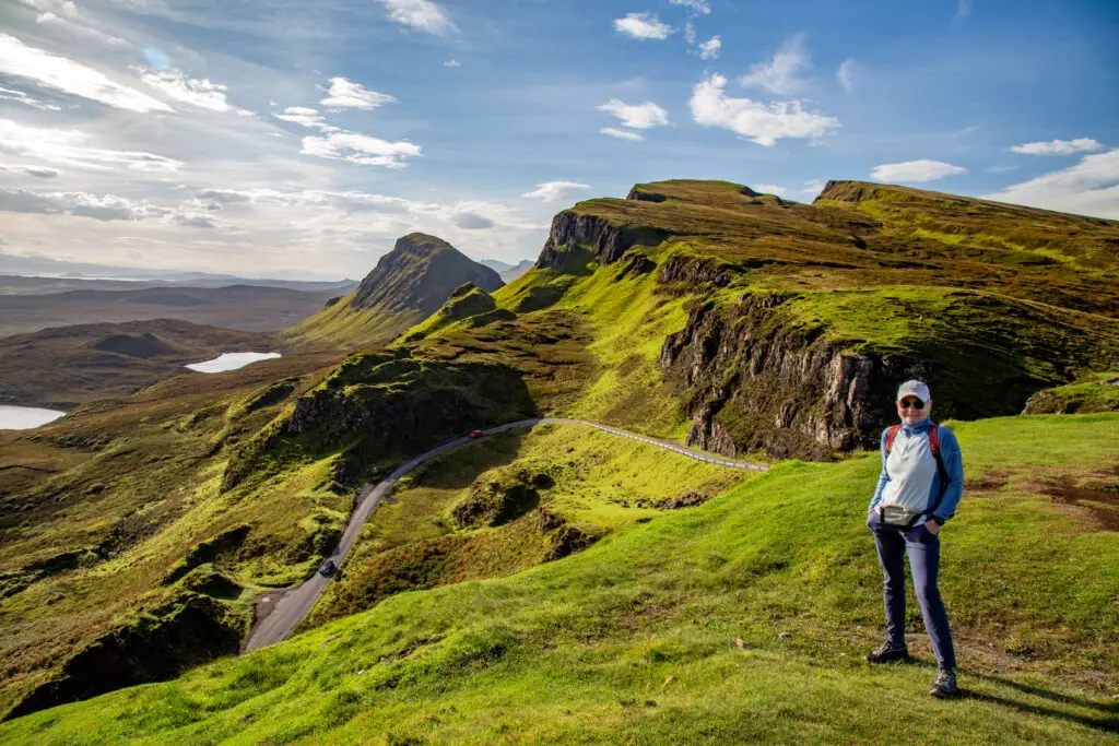 Tourist enjoying the scenic landscape of The Quiraing on the Isle of Skye, Scotland, with dramatic green hills, cliffs, and winding roads under a bright blue sky