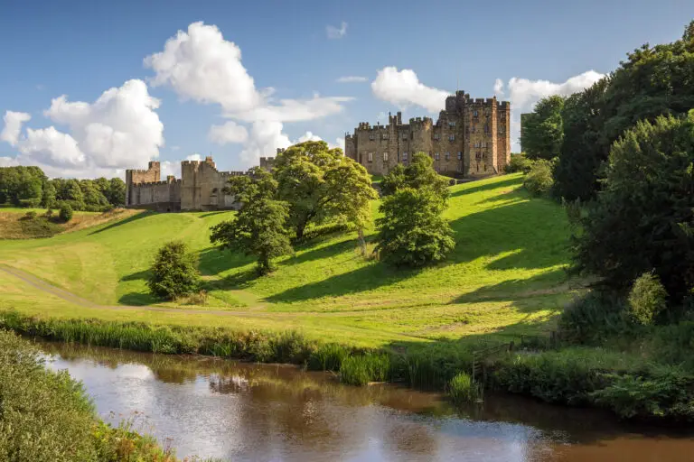 Alnwick Castle in England surrounded by lush green hills, trees, and a river under a bright blue sky, popular historic attraction featured in Braw Scottish Tours excursions.