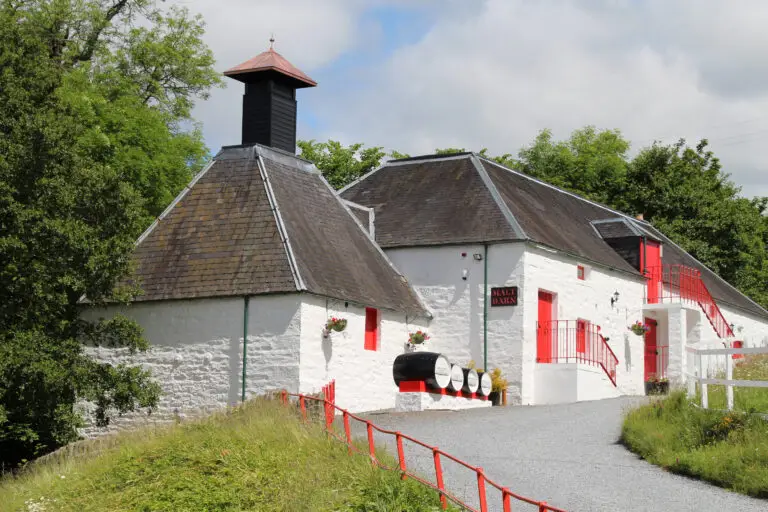 Traditional whitewashed distillery buildings with red doors and whisky barrels at Edradour Distillery in Pitlochry, Scotland, surrounded by greenery and blue skies
