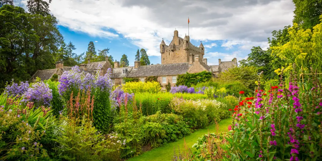 Cawdor Castle in Scotland surrounded by vibrant summer gardens and colorful flowers under a partly cloudy sky, popular tourist attraction on Braw Scottish Tours