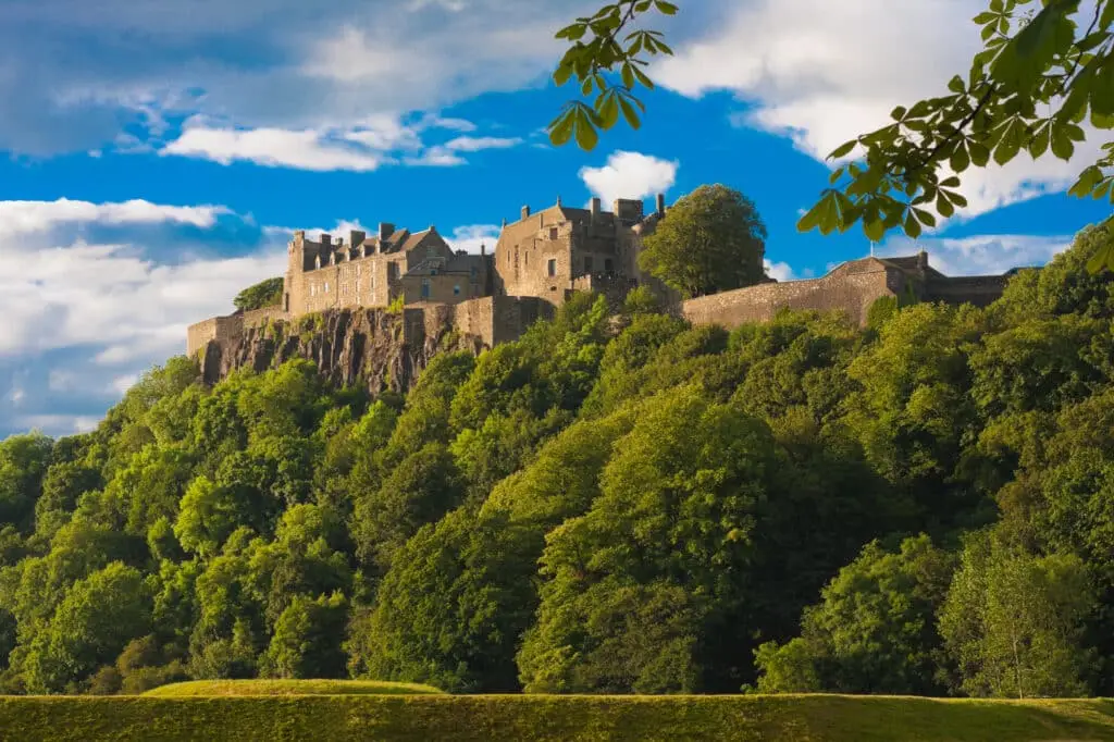Stirling Castle in Scotland perched on a rocky hill surrounded by lush green trees under a bright blue sky, a popular historic tourist attraction and landmark for Scottish tours