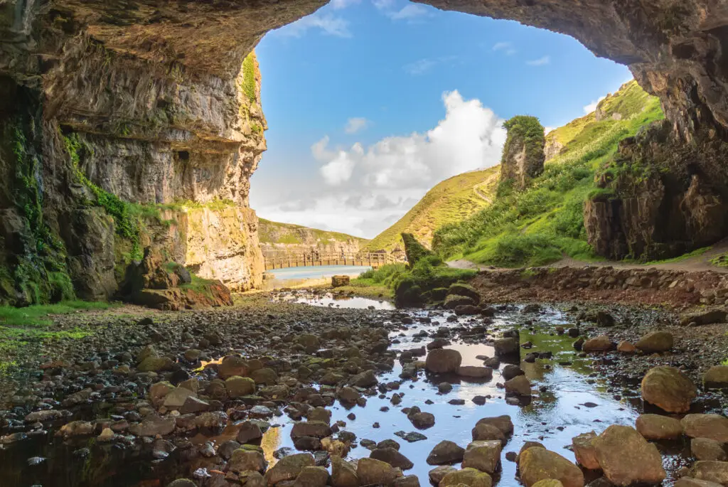 View from inside Smoo Cave in Durness, Scotland showing rocky cave walls, a stream leading to the sea, and green hills under a bright blue sky