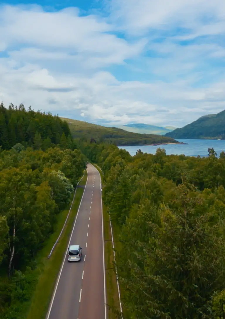 Luxury tour vehicle driving along scenic Loch Lomond road surrounded by lush green hills and blue water in Scotland