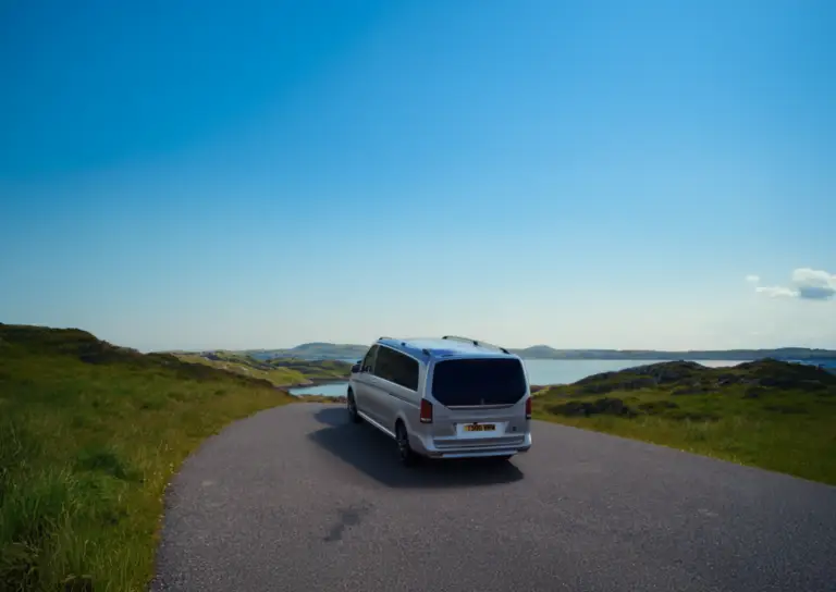 Luxury Mercedes tour vehicle driving along a scenic coastal road in Scotland with rolling green hills and sea views, part of Braw Scottish Tours experience