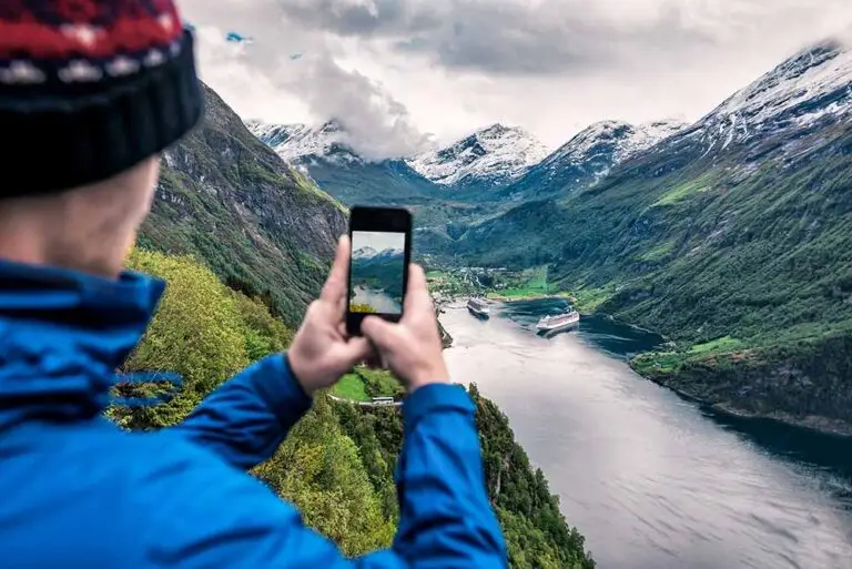 Tourist taking a photo of a scenic Norwegian fjord with cruise ships, snow-capped mountains, and lush green valleys in Norway